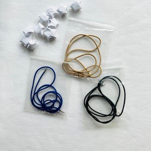 May include: An overhead shot shows three necklace cords in clear plastic bags, in tan, blue, and black. A collection of white origami stars are scattered above the bags. The cords are coiled and ready for use.