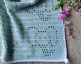 May include: A light mint green crochet blanket featuring a repeating heart pattern created with openwork stitches. The blanket has a solid white border along the edge and includes the text Crochessica DESIGNS printed in white script on the fabric.