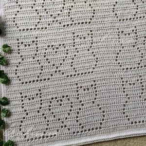 May include: A light grey crochet blanket featuring a repeating pattern of cat silhouettes created with openwork stitches. The rectangular textile has a solid border and a consistent grid-like texture throughout the entire surface of the fabric.