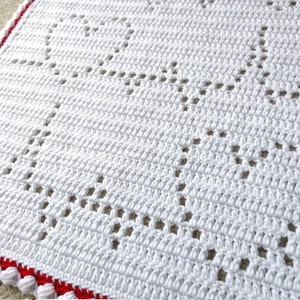 Op de afbeelding: Een witte gehaakte deken met een herhalend hartpatroon gemaakt met opengewerkte steken. De deken is afgewerkt met een levendige rode rand en een decoratieve witte bobbelrand, wat het ingewikkelde handwerk en de textuur benadrukt.