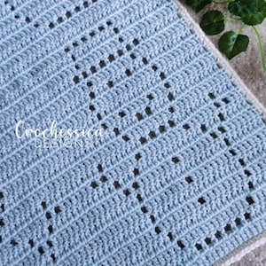 May include: A light blue crochet blanket with a white border and a repeating diamond pattern. The blanket has a crocheted texture and is made with a soft yarn. The text "Crochessica Designs" is visible in the lower left corner of the blanket.