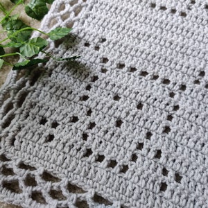 May include: A light grey crocheted blanket with a textured surface and decorative openwork border. The blanket features a repeating pattern of small, open diamond shapes. The overall design is simple and elegant.