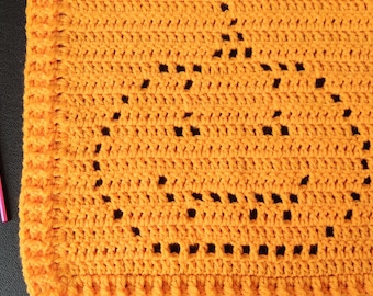 May include: A bright orange crocheted fabric featuring a pumpkin-shaped pattern created with openwork stitches. Beside the fabric lies a pink crochet hook and a small yellow plastic stitch marker, providing a sense of scale for the project.