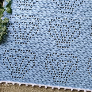 May include: A light blue crochet blanket featuring a repeating pattern of openwork hearts created with double crochet stitches. The blanket has a white scalloped border along the edge and is displayed next to a green leafy vine.