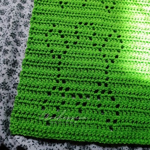 Może przedstawiać: Zielony pled szydełkowy z powtarzającym się wzorem małych otworów. Pled ma szydełkową obwódkę, a w prawym dolnym rogu widoczny jest tekst "Crochessica Designs".