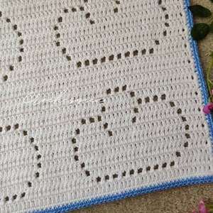 May include: A white crocheted blanket featuring a repeating heart pattern created with openwork stitches. The blanket has a light blue crocheted border along the edge. The text Crochessica DESIGNS is printed in white in the center of the blanket.