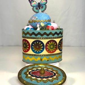 Può includere: Una scatola di latta rotonda vintage con coperchio, decorata con motivi floreali e geometrici colorati. La scatola è sormontata da un ornamento blu a forma di uovo, una farfalla e figurine in miniatura. Il coperchio è di lato.