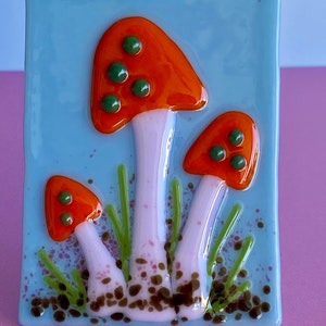 以下が含まれることがあります： 青い背景に、緑色の斑点のあるオレンジ色と白のキノコが3つ描かれたガラスのタイル。キノコは緑の草と茶色の土に囲まれています。