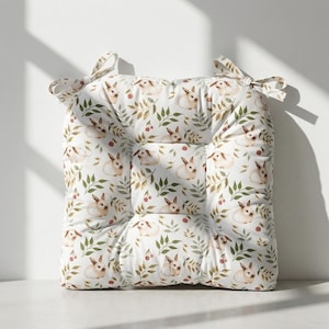 以下が含まれることがあります： 白い背景に、ウサギ、緑の葉、赤いベリーの繰り返し模様が施された正方形のシートクッション。 クッションはタフテッドデザインで、角に結び紐が付いています。
