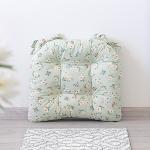 Könnte beinhalten: Ein quadratisches, gepolstertes Stuhlkissen mit Blumenmuster. Das Kissen ist hellgrün mit einem sich wiederholenden Muster aus rosa und weißen Blumen und kleinen blauen Akzenten. Es hat Bänder oben zum Befestigen an einem Stuhl.