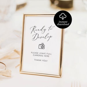 May include: A gold framed sign with the text "Ready to Develop" in elegant script. Below the text is a camera icon and the words "PLEASE LEAVE FULL CAMERAS HERE THANK YOU!" A black circle with a cloud and arrow icon is in the upper right corner.