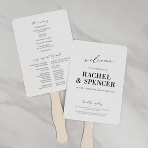 May include: Two white wedding ceremony fans with black text. The left fan lists the order of the ceremony and the names of the wedding party. The right fan says "Welcome to the wedding of Rachel & Spencer" with the date and location. The fans have wooden handles.