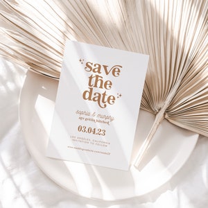 May include: A white "Save the Date" card with gold text, featuring the names "Sophia & Murphy" and the date "03.04.23". The card is on a white plate, with a dried palm leaf in the background. Wedding stationery.