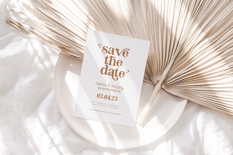 May include: A white "Save the Date" card with gold text, featuring the names "Sophia & Murphy" and the date "03.04.23". The card is on a white plate, with a dried palm leaf in the background. Wedding stationery.