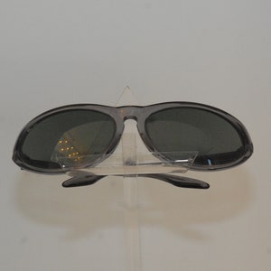 Pode incluir: Um par de &oacute;culos de sol ovais cinzentos com lentes escuras. Os &oacute;culos est&atilde;o em um suporte de pl&aacute;stico transparente.