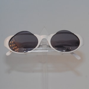 May include: White oval sunglasses with dark lenses. The sunglasses are on a clear stand.