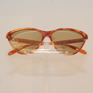 May include: A pair of tortoise shell sunglasses with brown lenses. The sunglasses are on a white display stand.