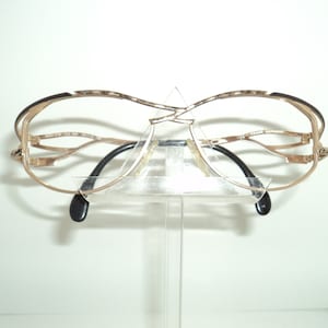 Op de afbeelding: Gouden metalen bril met een decoratief, ingewikkeld frame-ontwerp. De bril wordt getoond op een transparante kunststof standaard.