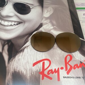 K&ouml;nnte beinhalten: Zwei runde, braun get&ouml;nte Ersatzgl&auml;ser f&uuml;r Sonnenbrillen mit dem Ray-Ban-Logo in der oberen Ecke des linken Glases. Die Gl&auml;ser liegen auf einer Werbebrosch&uuml;re, die eine Person mit einer Sonnenbrille zeigt.