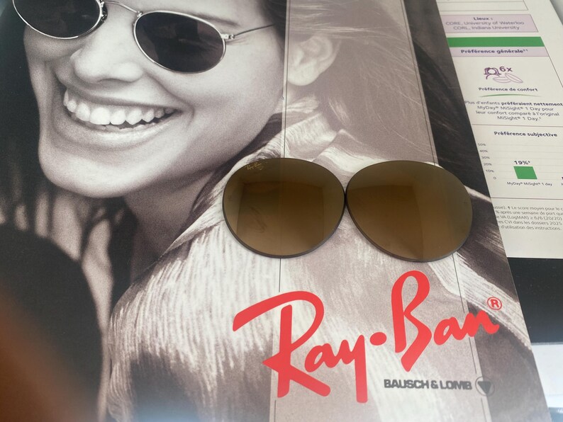 K&ouml;nnte beinhalten: Zwei runde, braun get&ouml;nte Ersatzgl&auml;ser f&uuml;r Sonnenbrillen mit dem Ray-Ban-Logo in der oberen Ecke des linken Glases. Die Gl&auml;ser liegen auf einer Werbebrosch&uuml;re, die eine Person mit einer Sonnenbrille zeigt.