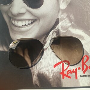 K&ouml;nnte beinhalten: Ein Paar dunkel get&ouml;nte Ray-Ban Sonnenbrillen mit goldfarbenem Logo an der oberen Ecke des linken Glases. Die Sonnenbrille liegt auf einer schwarz-wei&szlig;en Werbeanzeige, die das rote Ray-Ban Bausch & Lomb Logo deutlich zeigt.