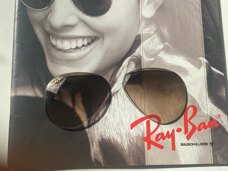 K&ouml;nnte beinhalten: Ein Paar dunkel get&ouml;nte Ray-Ban Sonnenbrillen mit goldfarbenem Logo an der oberen Ecke des linken Glases. Die Sonnenbrille liegt auf einer schwarz-wei&szlig;en Werbeanzeige, die das rote Ray-Ban Bausch & Lomb Logo deutlich zeigt.