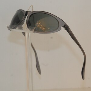 Pode incluir: &Oacute;culos de sol cinzentos com lentes ovais e arma&ccedil;&atilde;o fina. Os &oacute;culos de sol est&atilde;o em um suporte de pl&aacute;stico transparente.