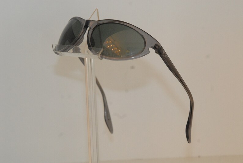 Pode incluir: &Oacute;culos de sol cinzentos com lentes ovais e arma&ccedil;&atilde;o fina. Os &oacute;culos de sol est&atilde;o em um suporte de pl&aacute;stico transparente.