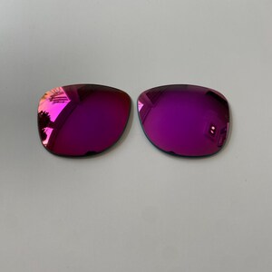 K&ouml;nnte beinhalten: Zwei Ersatz-Sonnenbrillengl&auml;ser mit einer leuchtenden, reflektierenden magenta- und lilafarbenen Spiegelbeschichtung. Die Gl&auml;ser haben eine abgerundete rechteckige Form und eine glatte, polierte Oberfl&auml;che f&uuml;r Brillengestelle.