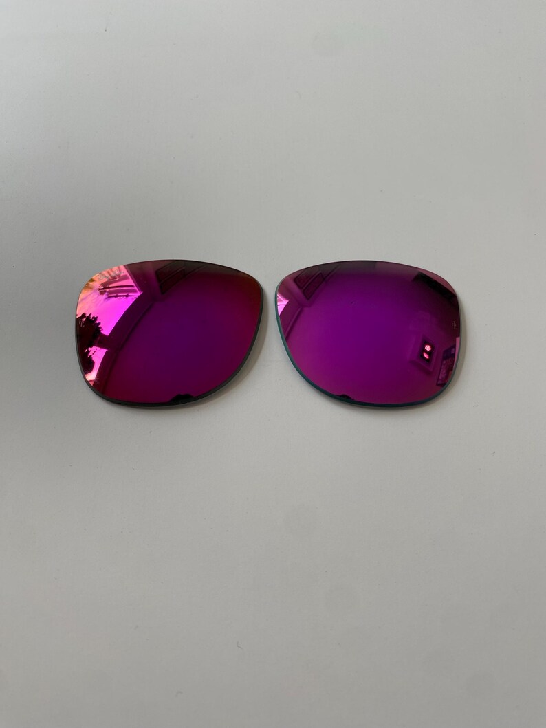 K&ouml;nnte beinhalten: Zwei Ersatz-Sonnenbrillengl&auml;ser mit einer leuchtenden, reflektierenden magenta- und lilafarbenen Spiegelbeschichtung. Die Gl&auml;ser haben eine abgerundete rechteckige Form und eine glatte, polierte Oberfl&auml;che f&uuml;r Brillengestelle.