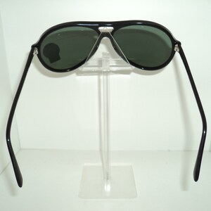 May include: Black aviator sunglasses with green lenses. The sunglasses are on a clear plastic stand.