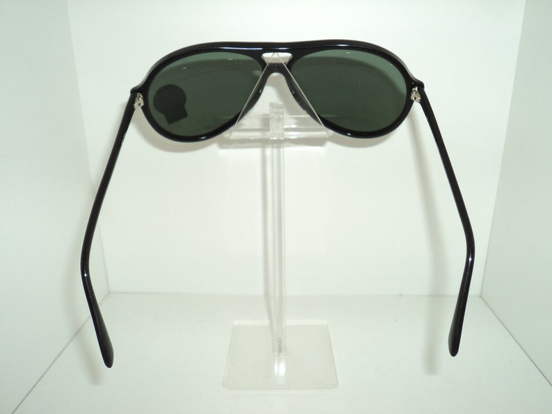 May include: Black aviator sunglasses with green lenses. The sunglasses are on a clear plastic stand.