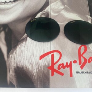 K&ouml;nnte beinhalten: Zwei dunkelgr&uuml;ne, kreisf&ouml;rmige Ersatzgl&auml;ser f&uuml;r Sonnenbrillen mit dem Ray-Ban-Logo in goldener Schrift in der oberen Ecke jedes Glases. Die Gl&auml;ser liegen auf einer gedruckten Anzeige mit dem roten Ray-Ban Bausch & Lomb-Logo.