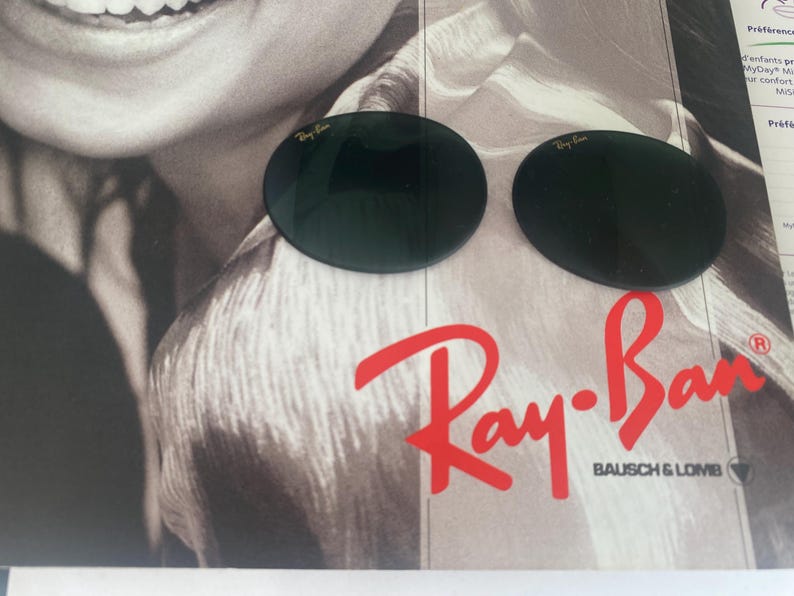 K&ouml;nnte beinhalten: Zwei dunkelgr&uuml;ne, kreisf&ouml;rmige Ersatzgl&auml;ser f&uuml;r Sonnenbrillen mit dem Ray-Ban-Logo in goldener Schrift in der oberen Ecke jedes Glases. Die Gl&auml;ser liegen auf einer gedruckten Anzeige mit dem roten Ray-Ban Bausch & Lomb-Logo.