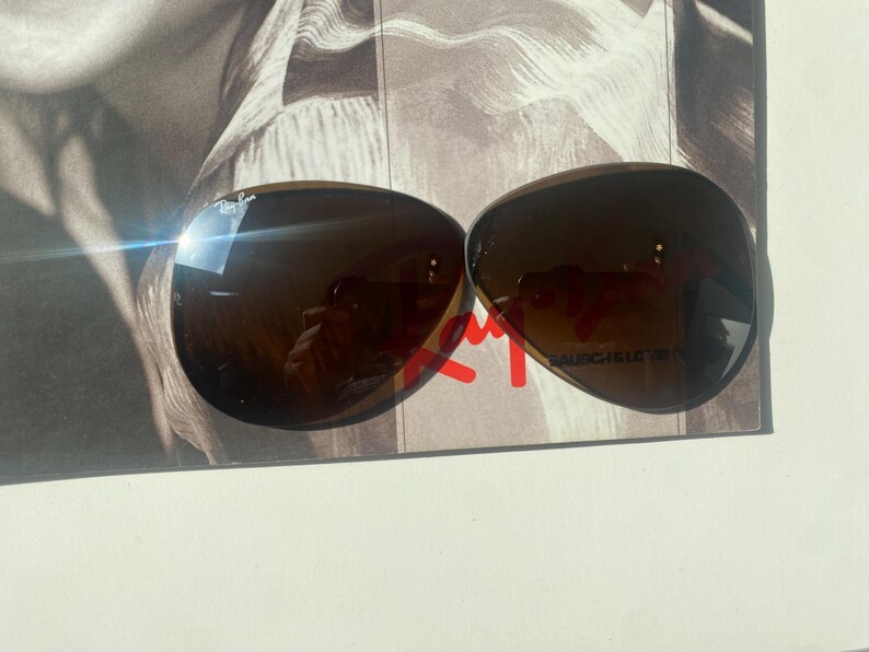 K&ouml;nnte beinhalten: Eine braun get&ouml;nte Fliegerbrille mit dem Ray-Ban-Logo auf dem oberen linken Glas und dem Schriftzug Bausch & Lomb auf dem unteren rechten Glas. Die tropfenf&ouml;rmigen Gl&auml;ser haben eine dunkle Verlaufsoptik und eine reflektierende Oberfl&auml;che.