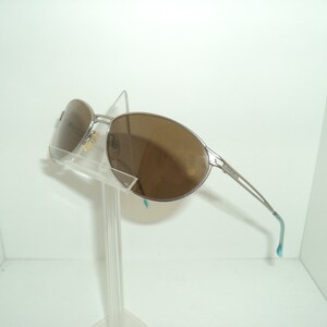 Puede incluir: Gafas de sol aviador plateadas con lentes marrones. Las gafas est&aacute;n en un expositor transparente.