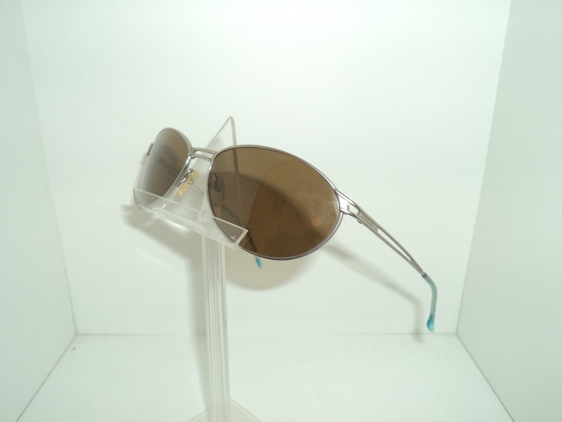 Puede incluir: Gafas de sol aviador plateadas con lentes marrones. Las gafas est&aacute;n en un expositor transparente.