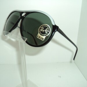 May include: Black Ray-Ban aviator sunglasses with green lenses and a white stripe on the frame. The sunglasses have a sticker on the frame that says "Ray-Ban 100% UV Protection G-15 Lens".
