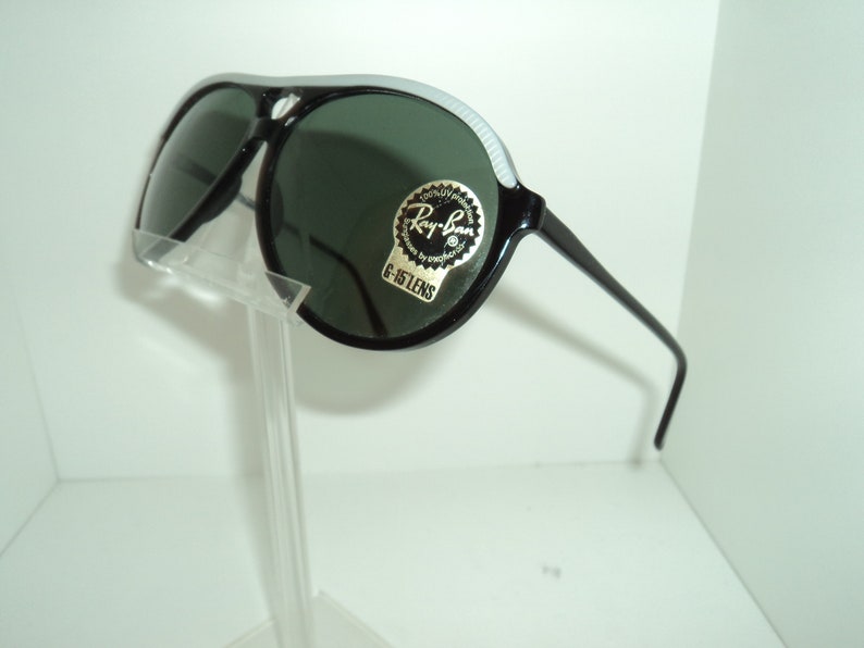 May include: Black Ray-Ban aviator sunglasses with green lenses and a white stripe on the frame. The sunglasses have a sticker on the frame that says "Ray-Ban 100% UV Protection G-15 Lens".