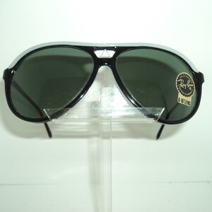 May include: Black aviator sunglasses with green lenses and a white stripe on the frame. The sunglasses have a Ray-Ban label with the text "G-15 LENS".