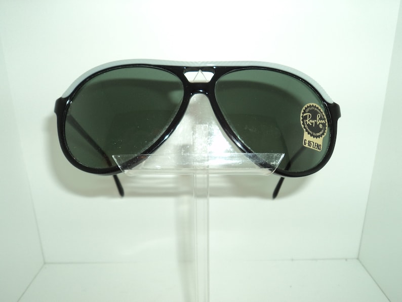 May include: Black aviator sunglasses with green lenses and a white stripe on the frame. The sunglasses have a Ray-Ban label with the text "G-15 LENS".