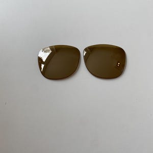 K&ouml;nnte beinhalten: Zwei separate, abgerundete quadratische Brillengl&auml;ser mit einer dunkelbraunen, reflektierenden T&ouml;nung. Die Gl&auml;ser haben glatte, geschwungene Kanten und eine polierte Oberfl&auml;che und sind so konzipiert, dass sie in einen Rahmen passen.