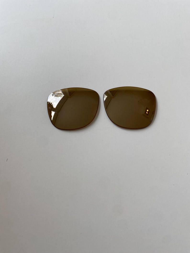 K&ouml;nnte beinhalten: Zwei separate, abgerundete quadratische Brillengl&auml;ser mit einer dunkelbraunen, reflektierenden T&ouml;nung. Die Gl&auml;ser haben glatte, geschwungene Kanten und eine polierte Oberfl&auml;che und sind so konzipiert, dass sie in einen Rahmen passen.