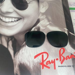 K&ouml;nnte beinhalten: Eine Sonnenbrille mit dunkel get&ouml;nten, abgerundeten Gl&auml;sern, die auf einer Werbeanzeige mit einem l&auml;chelnden Gesicht und dem roten Ray-Ban Bausch & Lomb Logo liegt. Die Sonnenbrille hat einen d&uuml;nnen Rahmen und dunkle, undurchsichtige Gl&auml;ser.