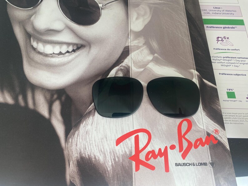 K&ouml;nnte beinhalten: Eine Sonnenbrille mit dunkel get&ouml;nten, abgerundeten Gl&auml;sern, die auf einer Werbeanzeige mit einem l&auml;chelnden Gesicht und dem roten Ray-Ban Bausch & Lomb Logo liegt. Die Sonnenbrille hat einen d&uuml;nnen Rahmen und dunkle, undurchsichtige Gl&auml;ser.
