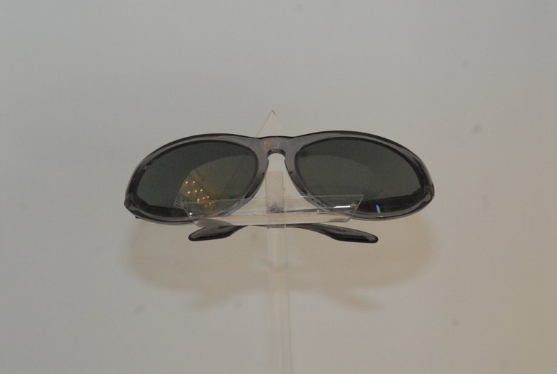 Pode incluir: Um par de &oacute;culos de sol ovais cinzentos com lentes escuras. Os &oacute;culos est&atilde;o em um suporte de pl&aacute;stico transparente.