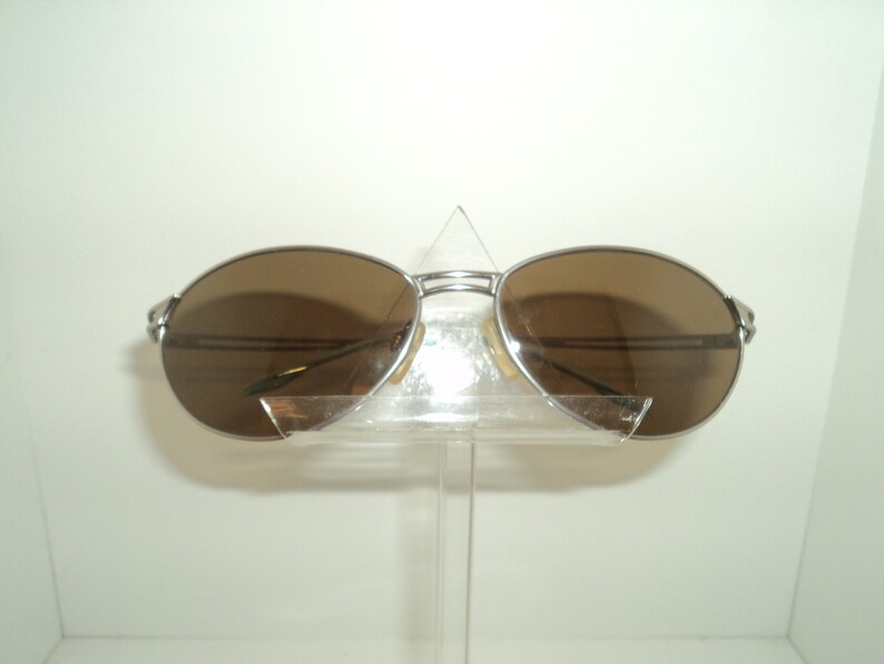 Puede incluir: Gafas de sol aviador plateadas con lentes marrones. Las gafas est&aacute;n en un soporte de pl&aacute;stico transparente.