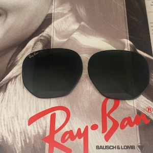 K&ouml;nnte beinhalten: Zwei dunkle, abgerundete Ersatz-Sonnenbrillengl&auml;ser mit dem goldfarbenen Ray-Ban-Logo in der oberen Ecke des linken Glases. Diese Gl&auml;ser sind f&uuml;r Bausch & Lomb-Rahmen konzipiert und werden vor einem markenbezogenen Werbehintergrund pr&auml;sentiert.