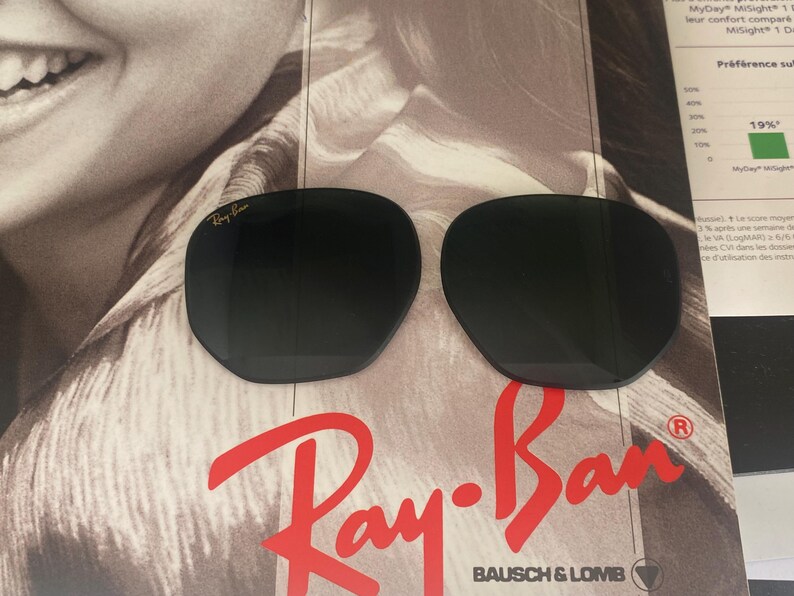 K&ouml;nnte beinhalten: Zwei dunkle, abgerundete Ersatz-Sonnenbrillengl&auml;ser mit dem goldfarbenen Ray-Ban-Logo in der oberen Ecke des linken Glases. Diese Gl&auml;ser sind f&uuml;r Bausch & Lomb-Rahmen konzipiert und werden vor einem markenbezogenen Werbehintergrund pr&auml;sentiert.