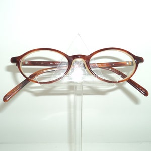 May include: Brown and beige oval-shaped eyeglasses with a clear plastic frame. The glasses are on a clear plastic stand.